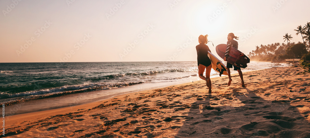 Young beautiful couple walking along the sandy beach near the ocean at sunset with surfboards, outdoor activities and sports holidays