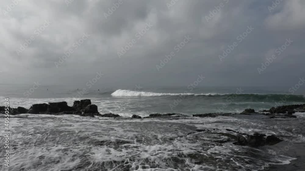 Waves crashing into the rocky shoreline of the pacific ocean while people are surfing in the background Waves crashing into the rocky shoreline of the pacific ocean while people are surfing in the background