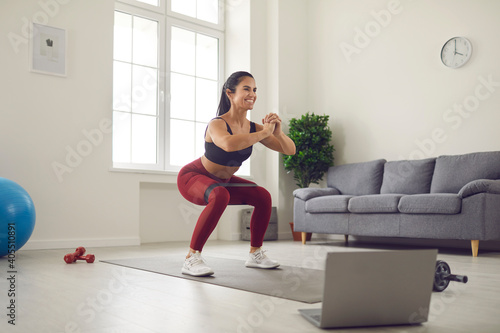 Fototapeta Naklejka Na Ścianę i Meble -  Smiling young woman working out at home with a rubber band in front of a laptop during an online broadcast of a fitness workout. Concept of activating the muscles of the buttocks and thighs.