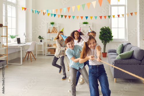Valokuva Team of happy smiling diverse children in funny cone hats playing tug-of-war in spacious decorated room at birthday party at home
