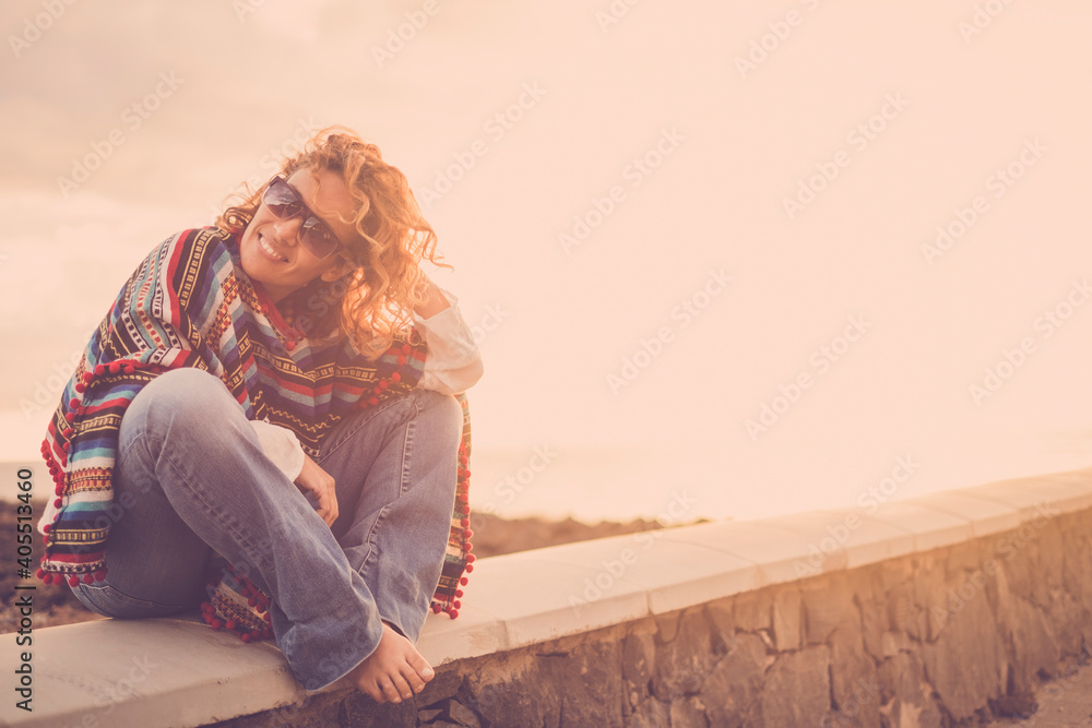Woman sit down outdoor enjoying sunset and freedom alone with a smile - portrait of cheerful female caucasian people with trendy clothes and mixeican style poncho - obrazy, fototapety, plakaty 