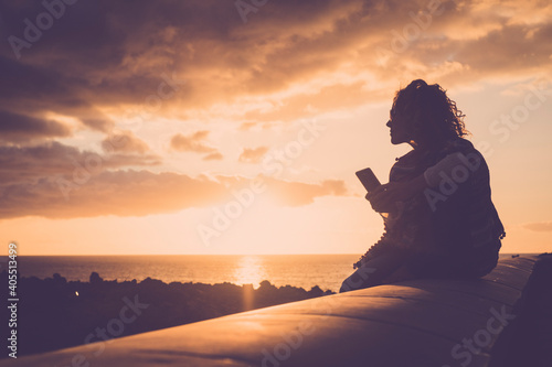 Tourist people enjoy sunset and beach on the coast sending message with a phone - woman sit down outdoor during evening looking ocean and using cellular smartphone alone - great sky in background photo