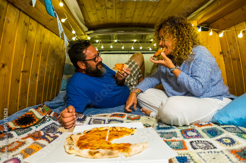 Couple have fun eating pizza inside vintage hand made wooden van vehicle in alternative lifestyle and travel vacation - happy romantic people enjoy holidays together in love