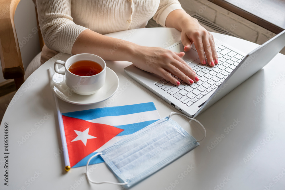 Woman tourist having breakfast with cup of coffee working on laptop. Flag of Cuba, medical protective face mask on table of cafe. Protection from bacteria and viruse in a public place. Concept. Woman tourist having breakfast with cup of coffee working on laptop. Flag of Cuba, medical protective face mask on table of cafe. Protection from bacteria and viruse in a public place. Concept.