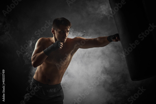 Aggressive boxer in black boxing wraps punching in boxing bag on dark background with smoke