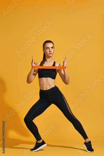 sporty beautiful woman in black sportswear exercising with rubber resistance band isolated on yellow background. Healthy lifestyle
