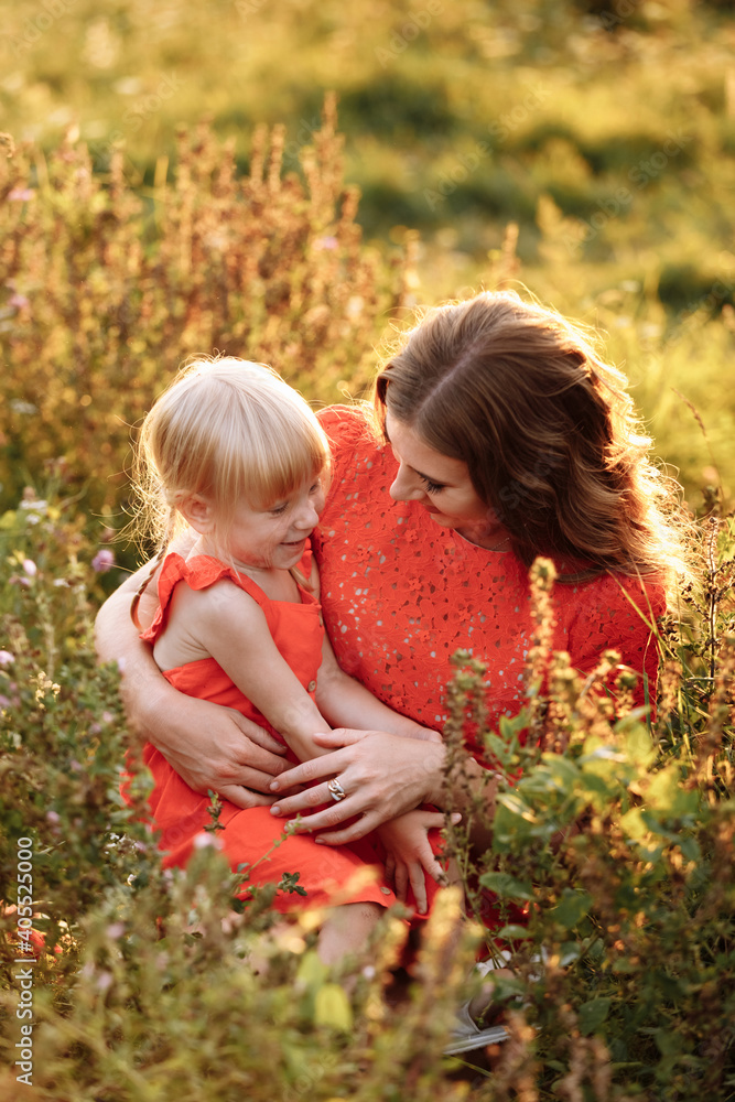 mothers day. womens day. Portrait of smiling beautiful young woman and her little daughter sitting on grass, against green of summer field on sunset. mothers day. womens day. Portrait of smiling beautiful young woman and her little daughter sitting on grass, against green of summer field on sunset.