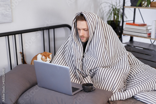 Feeling sick bearded attractive man in bed full covered with blanket using laptop and hot tea near him. Concept of distant work from home office or online studying during quarantine photo