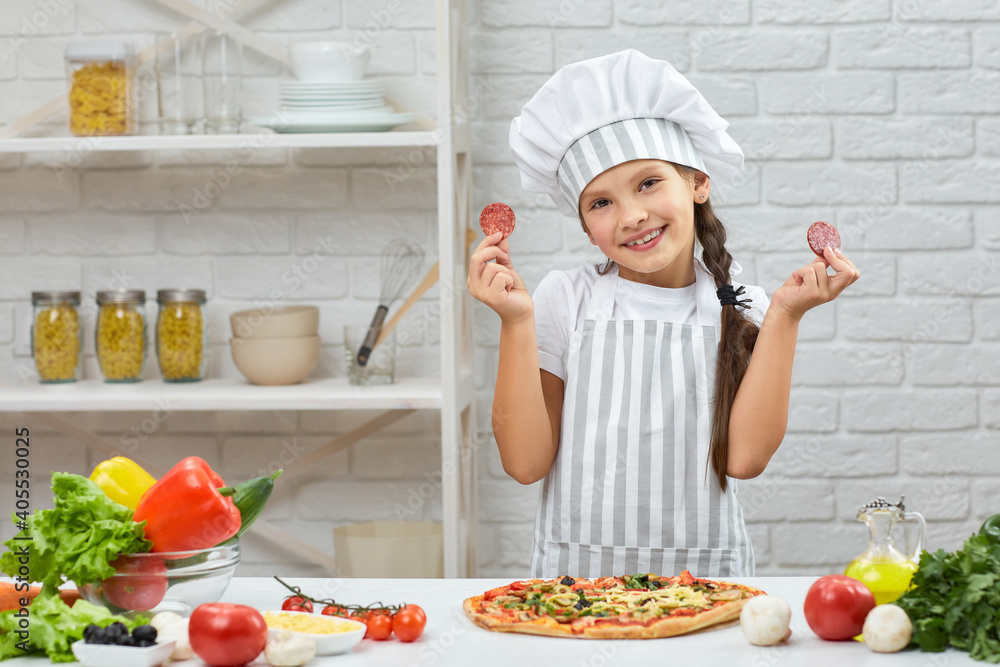 little girl in chef hat and an apron cooking pizza in the kitchen. the child holding sausage slices. having fun little girl in chef hat and an apron cooking pizza in the kitchen. the child holding sausage slices. having fun