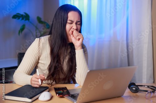 Young sleepy Asian woman sitting in front of computer yawning over worked at night, Asian female student study online at night