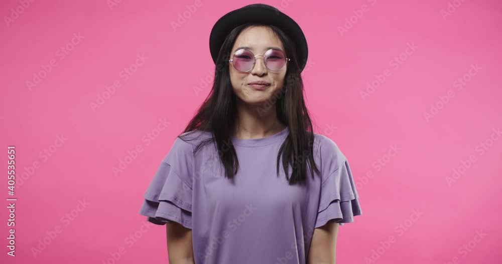 Close up portrait of happy cheerful young Asian female in modern glasses and black hat standing alone on pink background with smile on face looking at camera and smiling in good mood