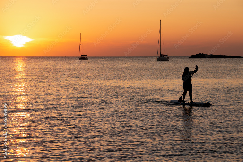 Man on stand up paddle board at sunset in calm waters with a sailboat