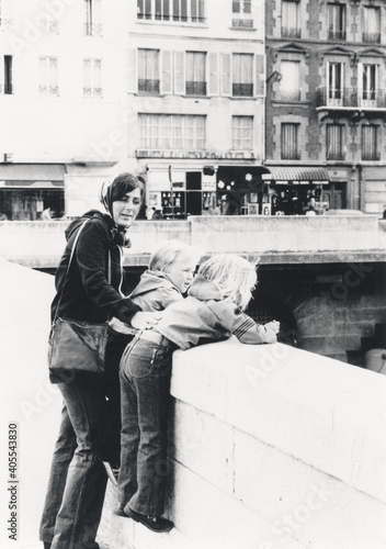Vintage retro 1978 monochrome image of a young mother in Paris with two children hanging over bridge railing over the Seine river, Paris, France.