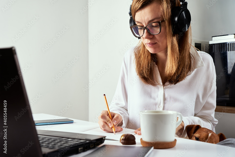 Remote work. Online courses, distance education and e learning concept. Woman in headphones listen audio course at laptop and makes notes in notebook Remote work. Online courses, distance education and e learning concept. Woman in headphones listen audio course at laptop and makes notes in notebook