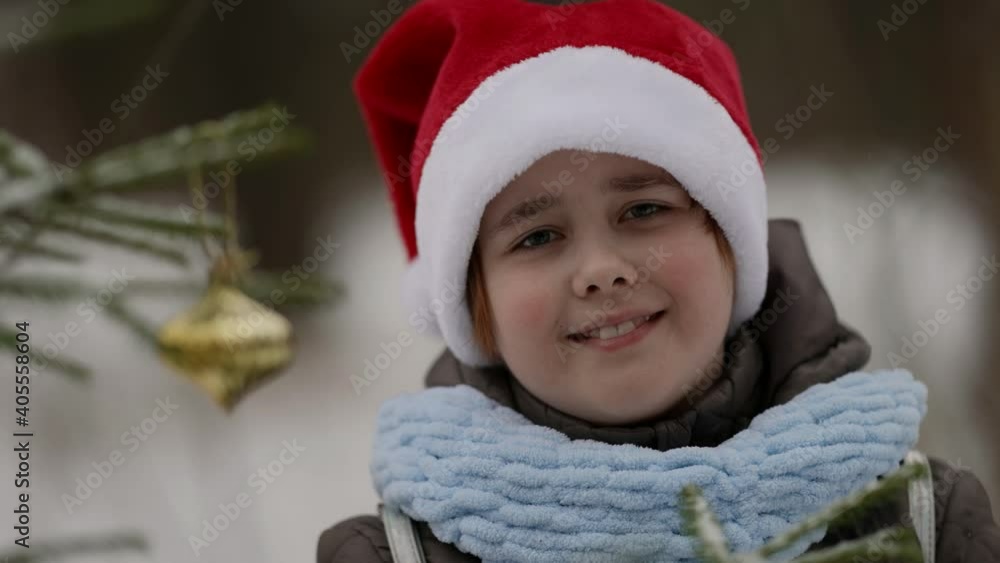 portrait of a small smiling girl in a Santa Claus hat and jacket, she walks outdoors on a winter day, stands near a street Christmas tree decorated with Christmas toys, snowy weather in the park. portrait of a small smiling girl in a Santa Claus hat and jacket, she walks outdoors on a winter day, stands near a street Christmas tree decorated with Christmas toys, snowy weather in the park.