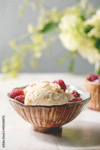 Homemade caramel vanilla ice cream with frozen raspberries in ceramic bowl standing on white marble table with flowers behind.