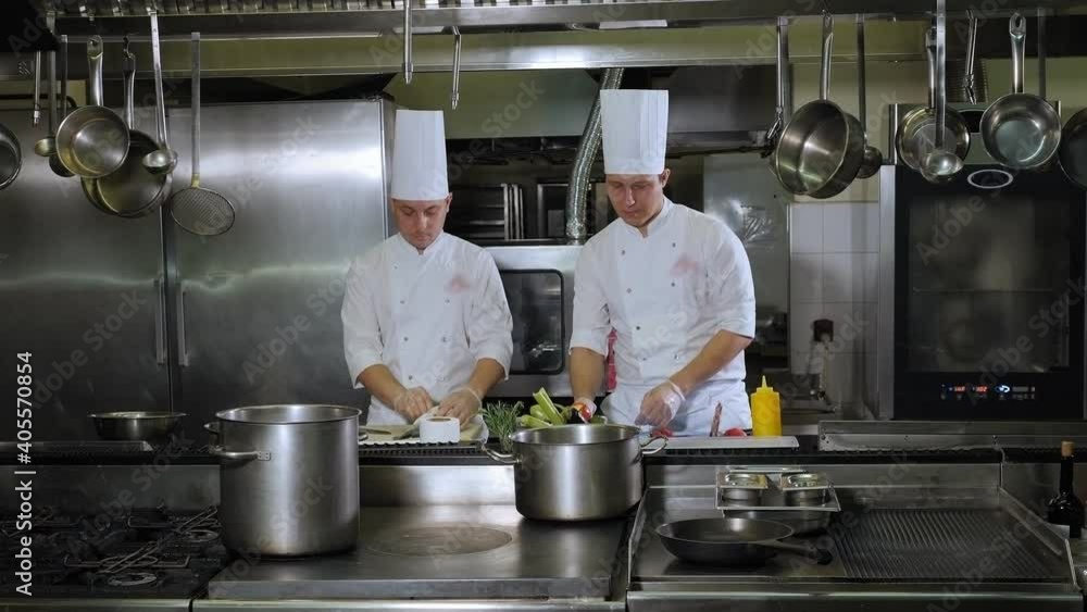 Two chefs in white uniforms work in a large professional kitchen, cutting vegetables and fish. Preparing dinner in a professional restaurant kitchen. Two chefs in white uniforms work in a large professional kitchen, cutting vegetables and fish. Preparing dinner in a professional restaurant kitchen.