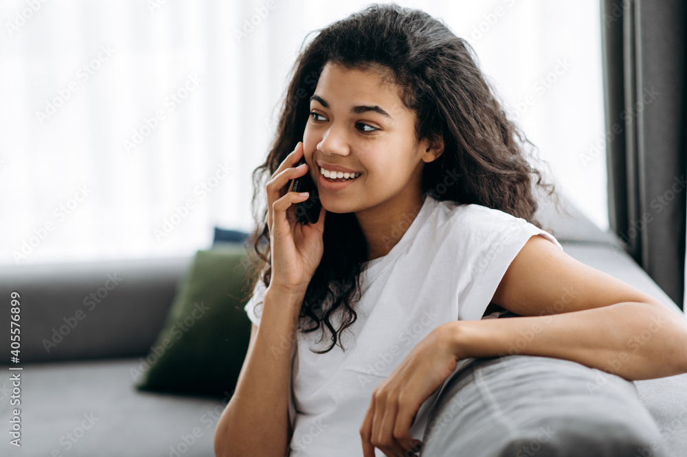 Pretty african american girl sits on the sofa at home. Attractive young woman talking by phone with friend, family or boyfriend, looks away and smiling. Leisure time at home Pretty african american girl sits on the sofa at home. Attractive young woman talking by phone with friend, family or boyfriend, looks away and smiling. Leisure time at home