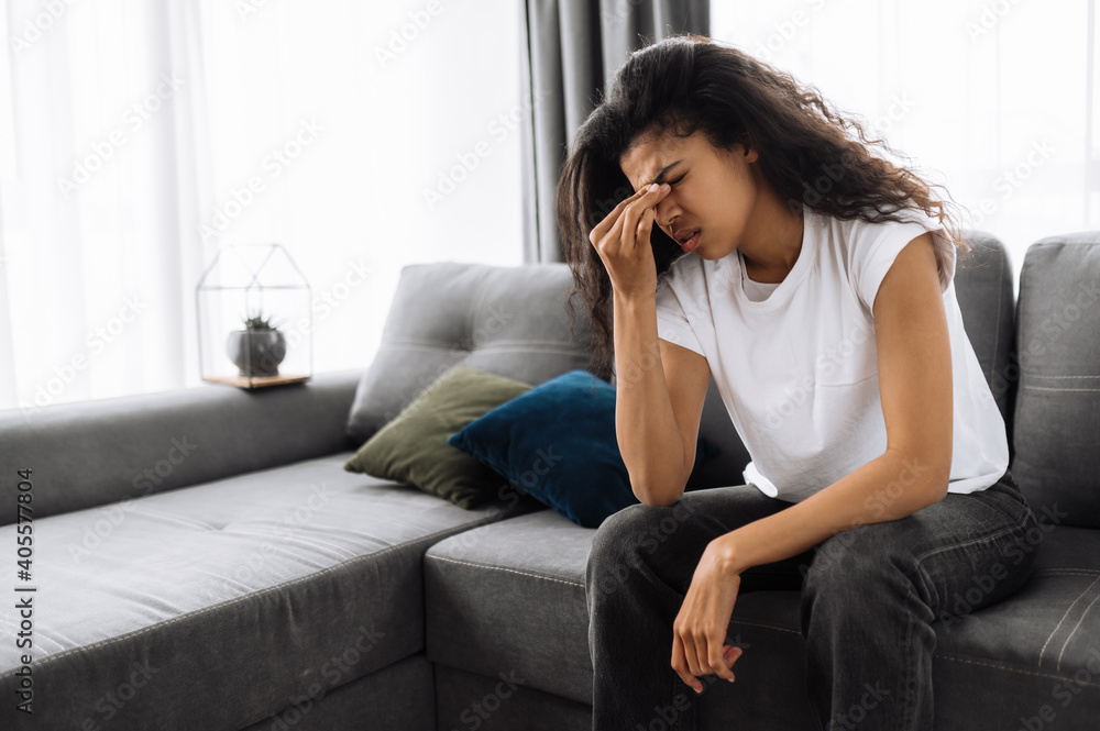 Tired girl sitting on the sofa at home, having a terrible headache. Exhausted african american female massage the eyes, feeling unhealthy. Young woman catch a cold or having flu, upset about illness Tired girl sitting on the sofa at home, having a terrible headache. Exhausted african american female massage the eyes, feeling unhealthy. Young woman catch a cold or having flu, upset about illness
