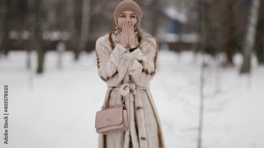 An attractive fashionable blonde in a beautiful fur coat and a beige hat poses against the background of a winter park. The model is outside on a snowy background, rubbing her hands against the cold.