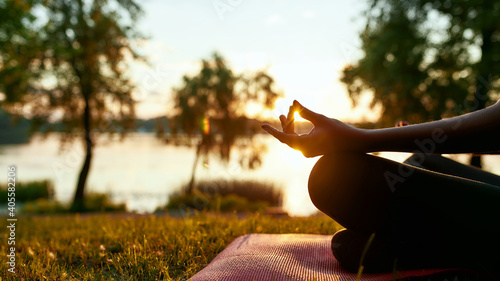 Cropped shot of a woman doing yoga outdoors near lake or river at sunrise in the morning, sitting in lotus pose on the background of nature