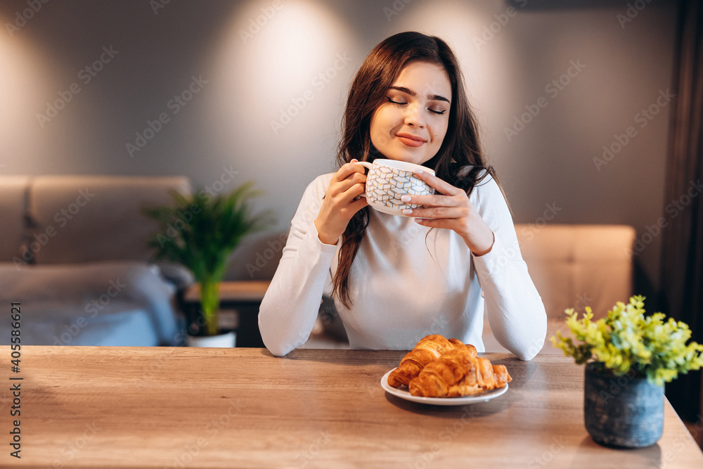 Young woman with black shiny hair drinking coffee during breakfast. Indoor portrait of cute brunette girl eating croissant and enjoying tea in morning Young woman with black shiny hair drinking coffee during breakfast. Indoor portrait of cute brunette girl eating croissant and enjoying tea in morning