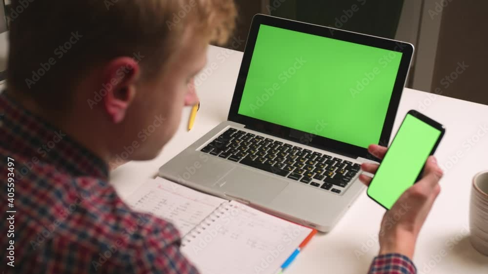 Back view. Close up man use laptop green screen holding phone chromakey at home office. Man watching content on phone green screen chromakey. Back view. Close up man use laptop green screen holding phone chromakey at home office. Man watching content on phone green screen chromakey.