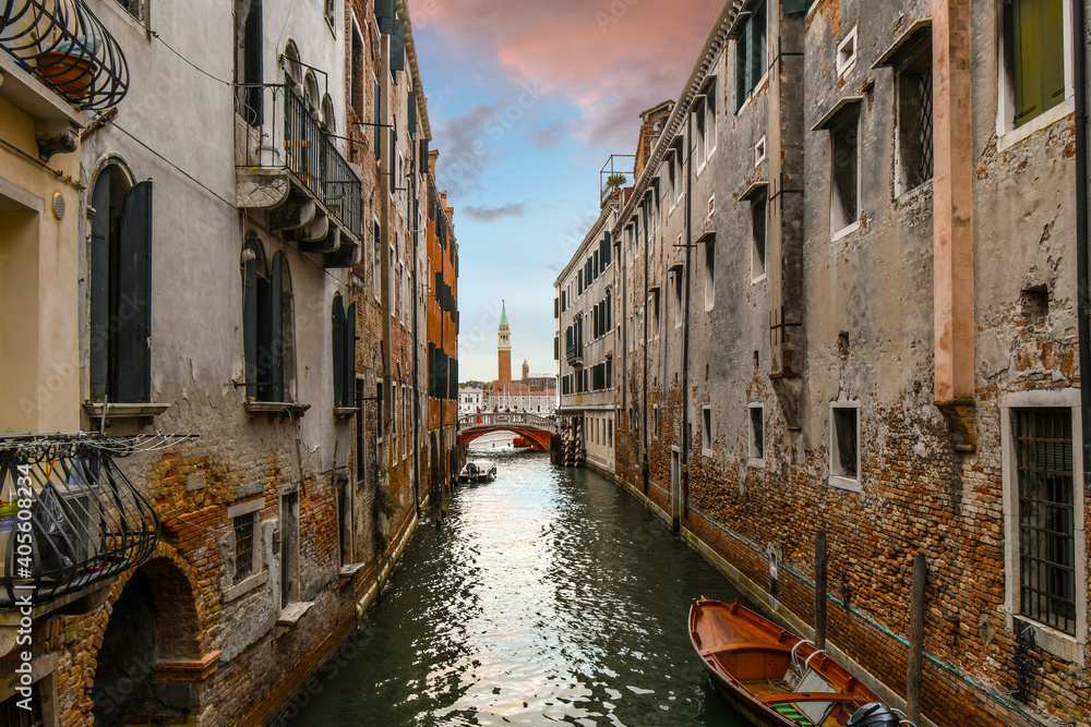 Tourists cross a bridge over a residential canal off the beaten path with a cathedral bell tower in view across the grand canal in Venice, Italy