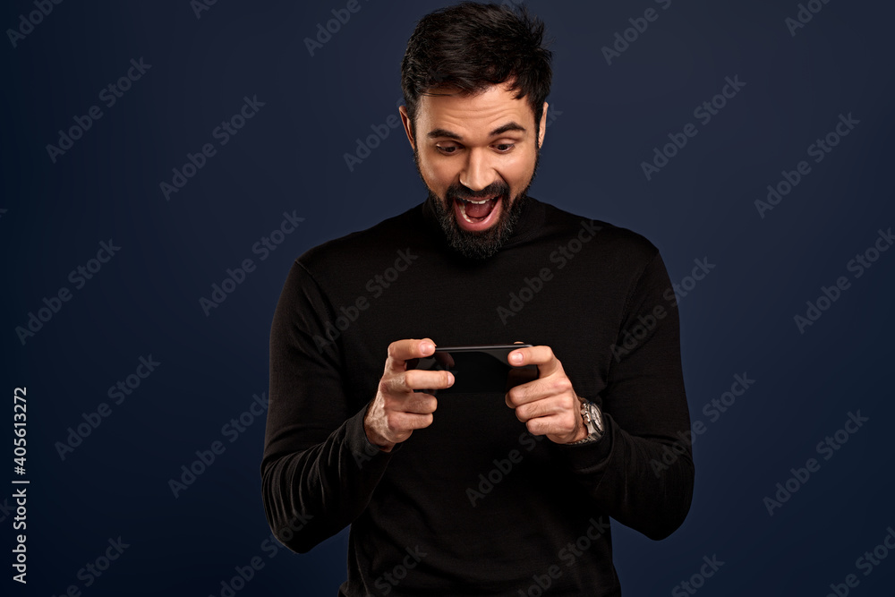 Portrait of excited, happy bearded man holding mobile phone horizontally and smiling amused at display, playing awesome new game, upload arcade, watching funny video, over Pacific Blue background Portrait of excited, happy bearded man holding mobile phone horizontally and smiling amused at display, playing awesome new game, upload arcade, watching funny video, over Pacific Blue background