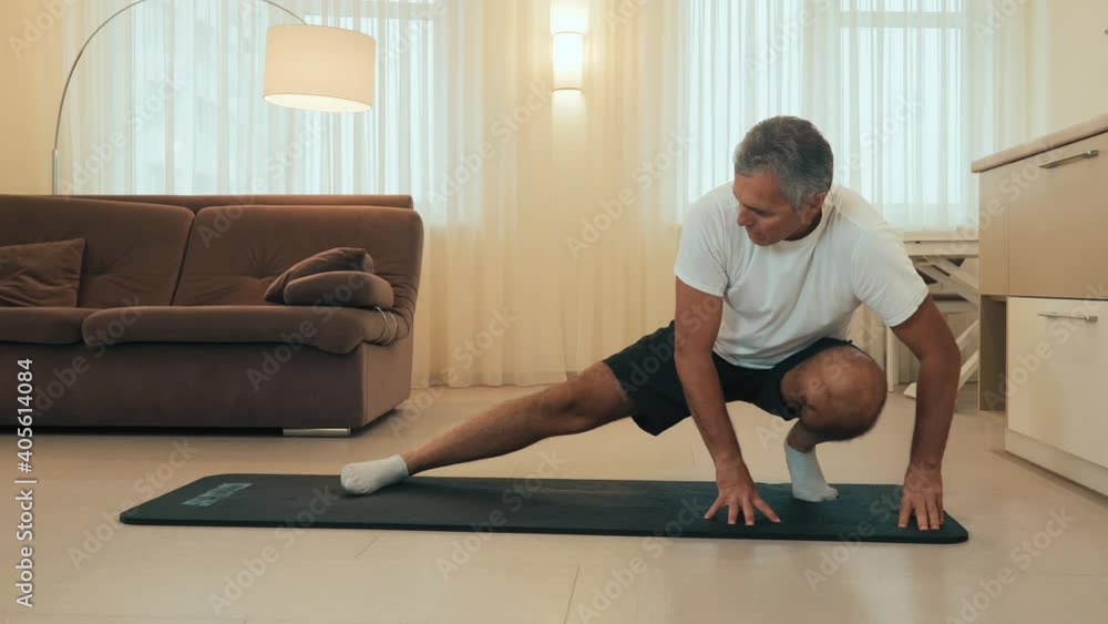Elderly man doing side lunge exercise. Senior male athlete with fit body warming up, doing leg stretch workout at home. Take care of yourself Modern light room on background. Elderly sport concept Elderly man doing side lunge exercise. Senior male athlete with fit body warming up, doing leg stretch workout at home. Take care of yourself Modern light room on background. Elderly sport concept