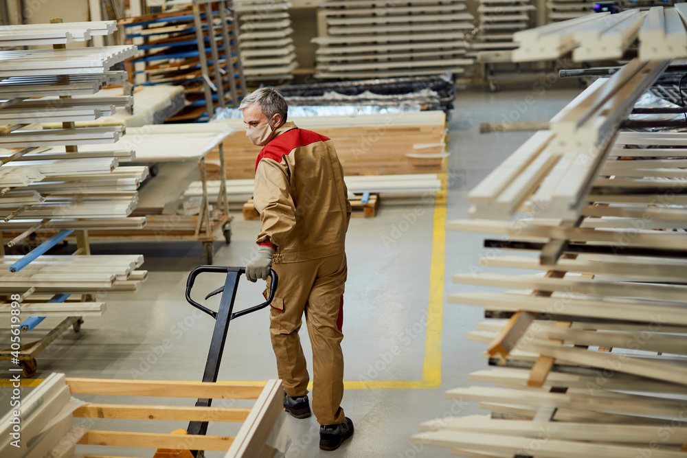 Fototapeta premium Full length of carpenter with face mask pulling pallet jack in a workshop.