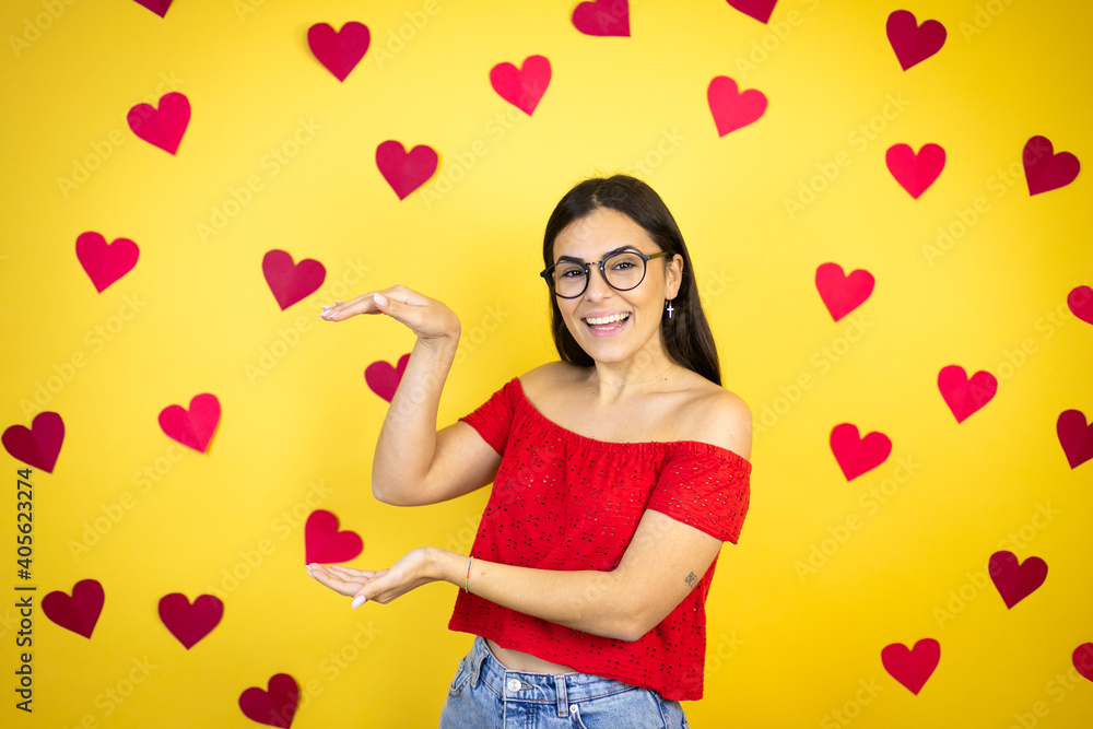 Young beautiful woman over yellow background with red hearts gesturing with hands showing big and large size sign, measure symbol. Smiling . Measuring concept. Young beautiful woman over yellow background with red hearts gesturing with hands showing big and large size sign, measure symbol. Smiling . Measuring concept.