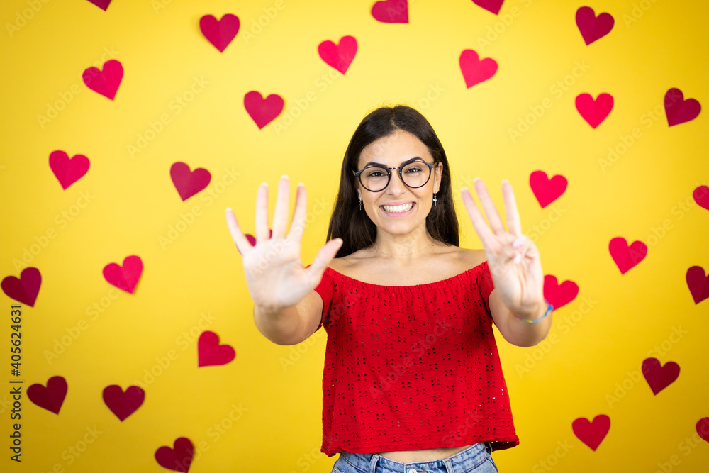 Young beautiful woman over yellow background with red hearts showing and pointing up with fingers number eight while smiling confident and happy Young beautiful woman over yellow background with red hearts showing and pointing up with fingers number eight while smiling confident and happy