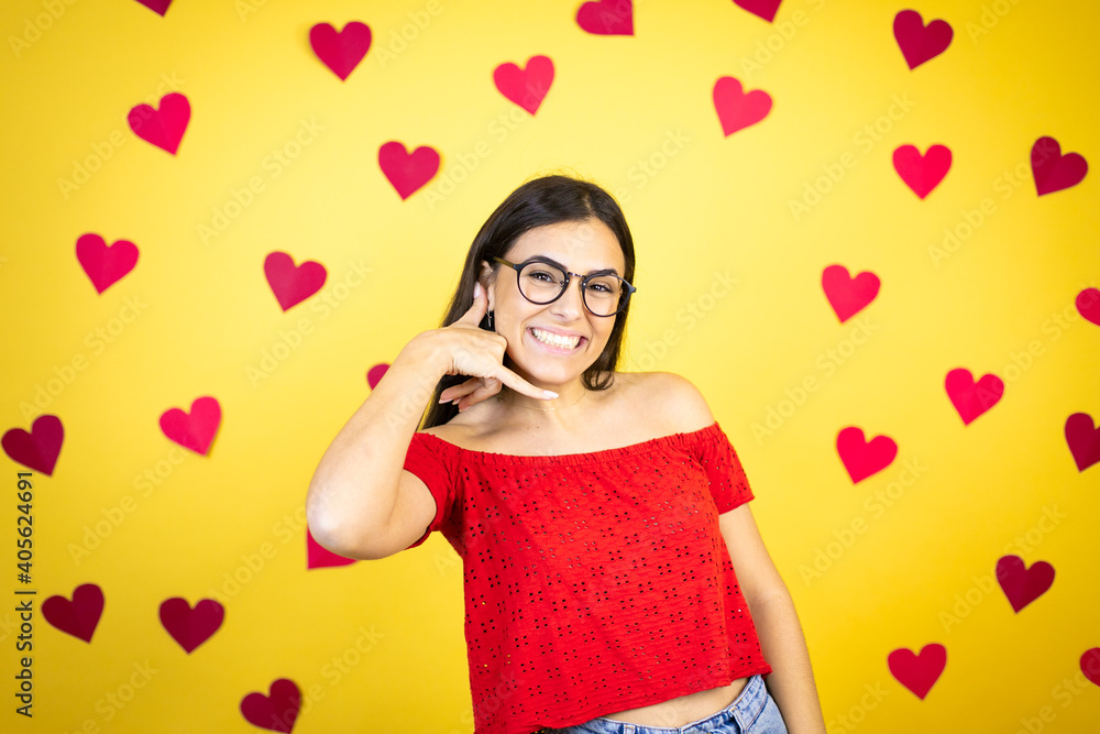 Young beautiful woman over yellow background with red hearts smiling doing phone gesture with hand and fingers like talking on the telephone Young beautiful woman over yellow background with red hearts smiling doing phone gesture with hand and fingers like talking on the telephone