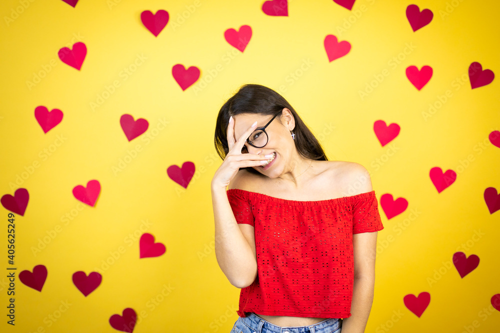 Young beautiful woman over yellow background with red hearts peeking in shock covering face and eyes with hand, looking through fingers with embarrassed expression Young beautiful woman over yellow background with red hearts peeking in shock covering face and eyes with hand, looking through fingers with embarrassed expression