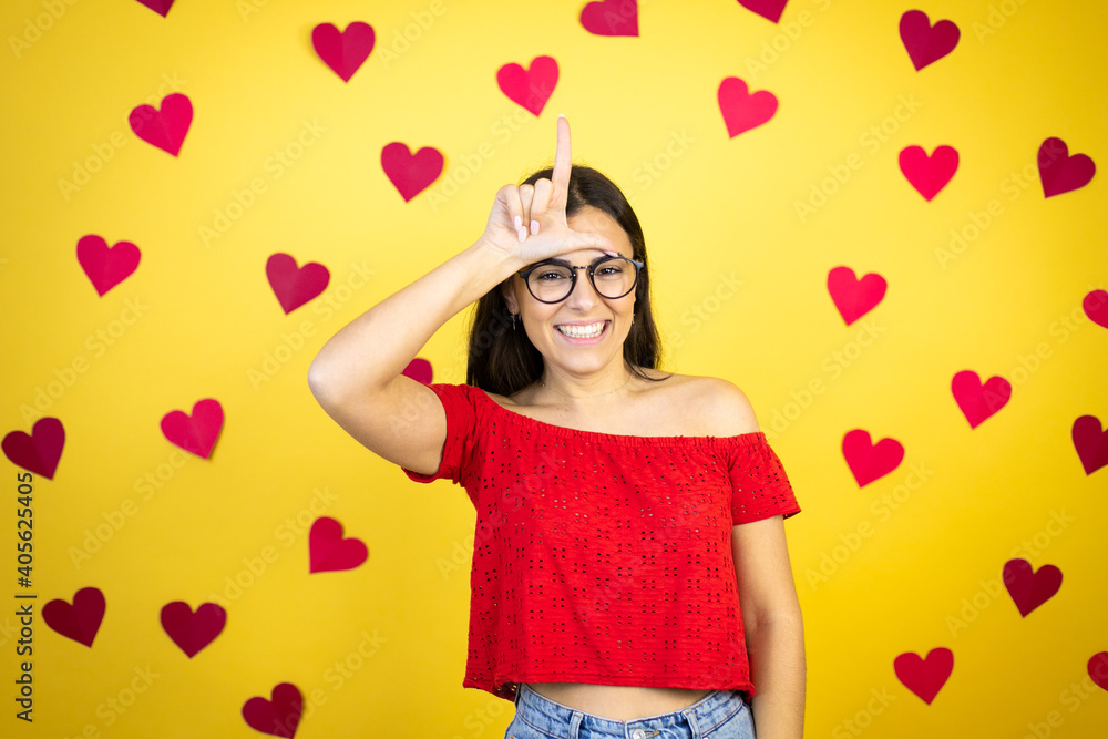 Young beautiful woman over yellow background with red hearts making fun of people with fingers on forehead doing loser gesture mocking and insulting. Young beautiful woman over yellow background with red hearts making fun of people with fingers on forehead doing loser gesture mocking and insulting.