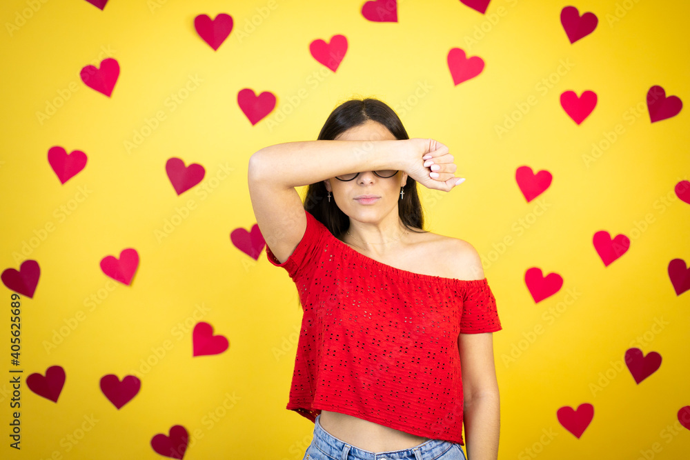 Young beautiful woman over yellow background with red hearts covering eyes with arm smiling cheerful and funny. Blind concept. Young beautiful woman over yellow background with red hearts covering eyes with arm smiling cheerful and funny. Blind concept.