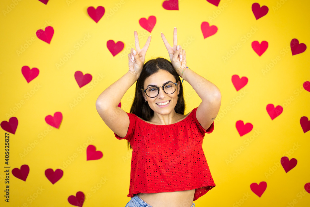 Young beautiful woman over yellow background with red hearts Posing funny and crazy with fingers on head as bunny ears, smiling cheerful Young beautiful woman over yellow background with red hearts Posing funny and crazy with fingers on head as bunny ears, smiling cheerful