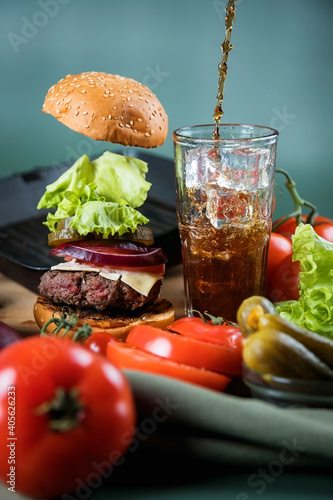 Classic burger and its ingredients. A dark carbonated drink is poured into a tall glass glass with ice.
