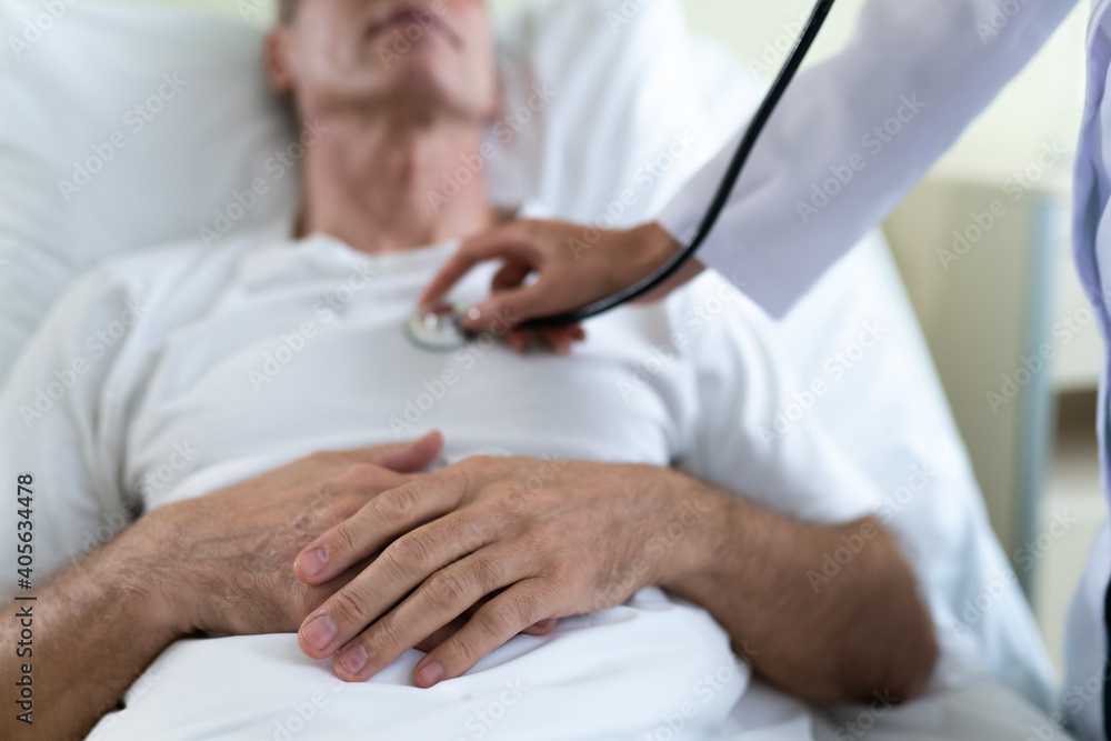 Young modern confident woman doctor in a white coat with a stethoscope is examining the patient's feelings while he is lying on the bed in the hospital ward. Young modern confident woman doctor in a white coat with a stethoscope is examining the patient's feelings while he is lying on the bed in the hospital ward.