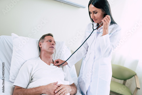 Young modern confident woman doctor in a white coat with a stethoscope is examining the patient s feelings while he is lying on the bed in the hospital ward.