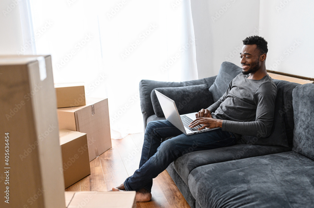 Successful freelancer using laptop, sit on the couch in living room at new apartment. Happy african american man searching ideas for interior design or work project. Male employee remotely working Successful freelancer using laptop, sit on the couch in living room at new apartment. Happy african american man searching ideas for interior design or work project. Male employee remotely working