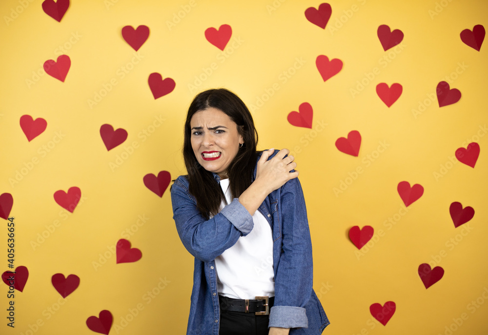 Young caucasian woman over yellow background with red hearts with pain on her shoulder and a painful expression Young caucasian woman over yellow background with red hearts with pain on her shoulder and a painful expression