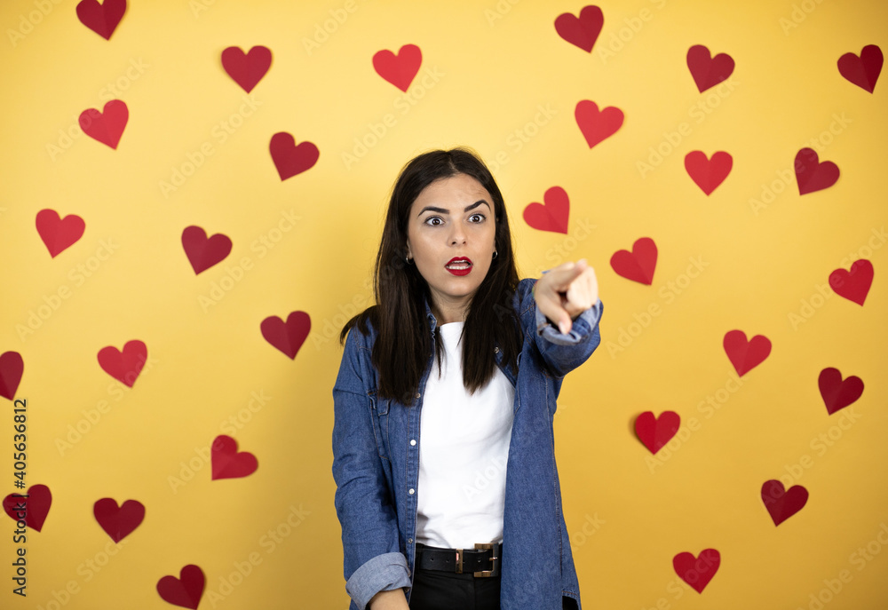 Young caucasian woman over yellow background with red hearts pointing with finger surprised ahead, open mouth amazed expression, something on the front Young caucasian woman over yellow background with red hearts pointing with finger surprised ahead, open mouth amazed expression, something on the front
