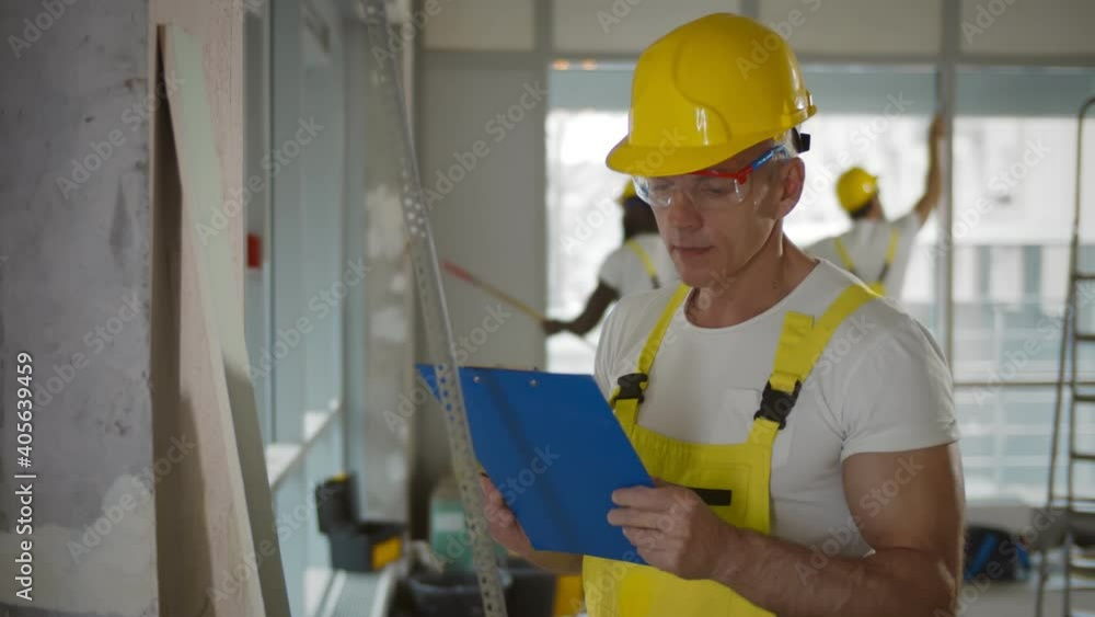 Foreman in uniform holding clipboard and making marks on wall in renovating room Foreman in uniform holding clipboard and making marks on wall in renovating room