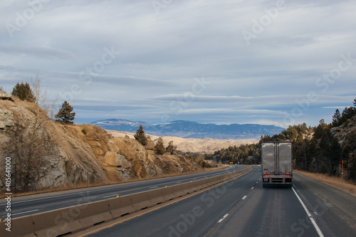 A road landscape on an autumn day, a highway on the sides of which there are sheer rocks with large stones, among which trees grow, trucks are driving, a beautiful sky, mountains in the distance. © Liudmila