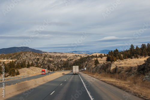 A road landscape on an autumn day, a highway on the sides of which there are sheer rocks with large stones, among which trees grow, trucks are driving, a beautiful sky, mountains in the distance.