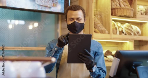 Young African American bakery worker stands on background of shelves with bread in protective mask and makes ordering products online using tablet. photo