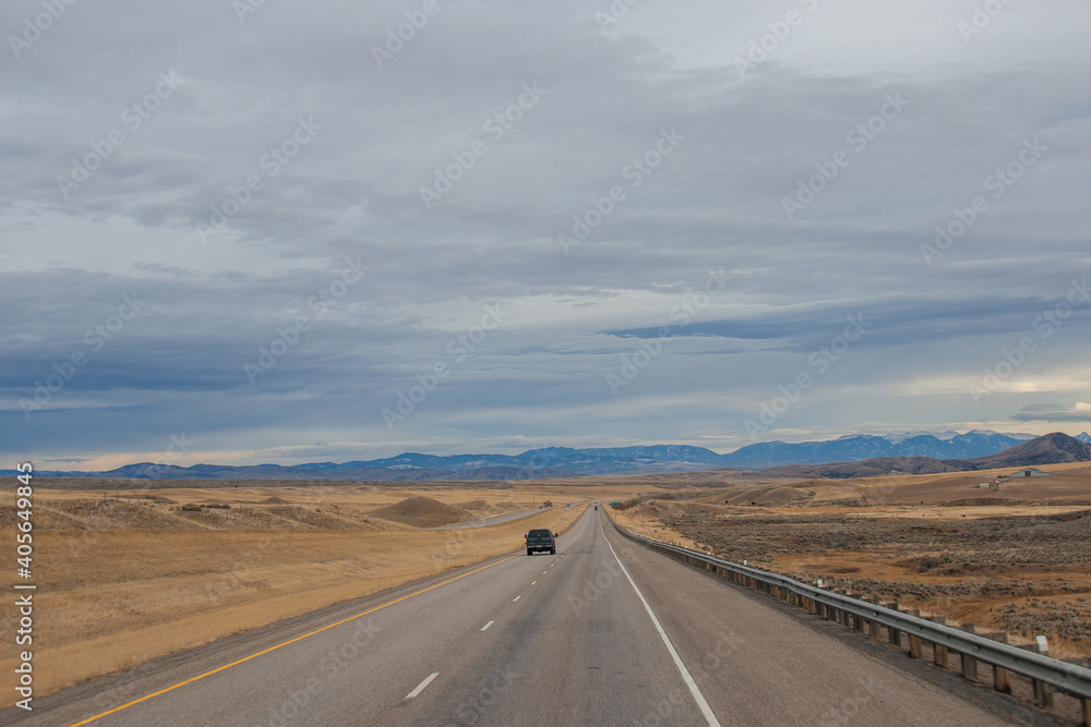 Asphalt road among steppes with yellow dry grass, blue mountains on the horizon, beautiful blue-gray autumn sky. Montana, USA, 11-23-2019