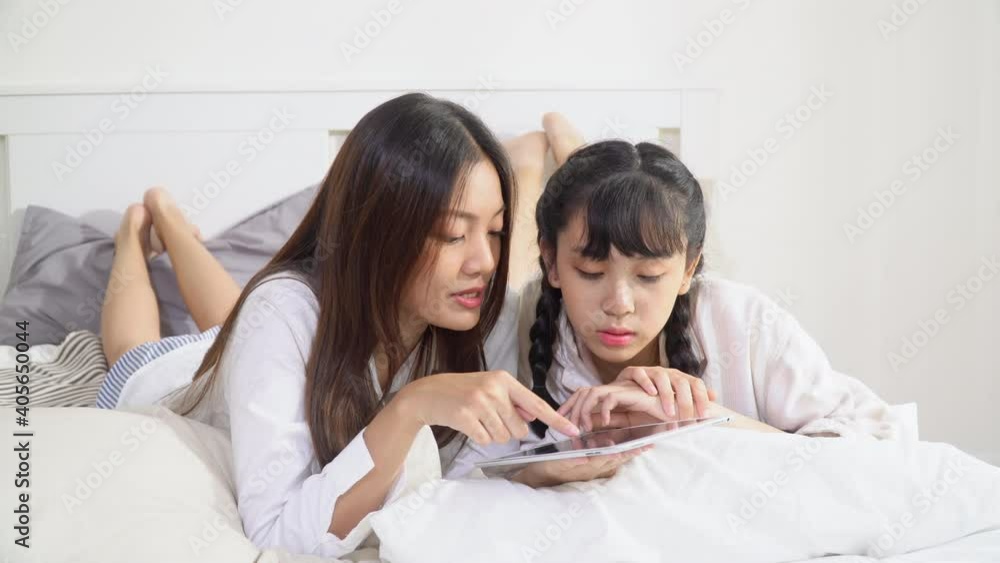 Smiling young beautiful Asian woman lying down on the bed and teaching her sister for learning education with using digital tablet and internet. Home study, happy family and technology concept. Smiling young beautiful Asian woman lying down on the bed and teaching her sister for learning education with using digital tablet and internet. Home study, happy family and technology concept.